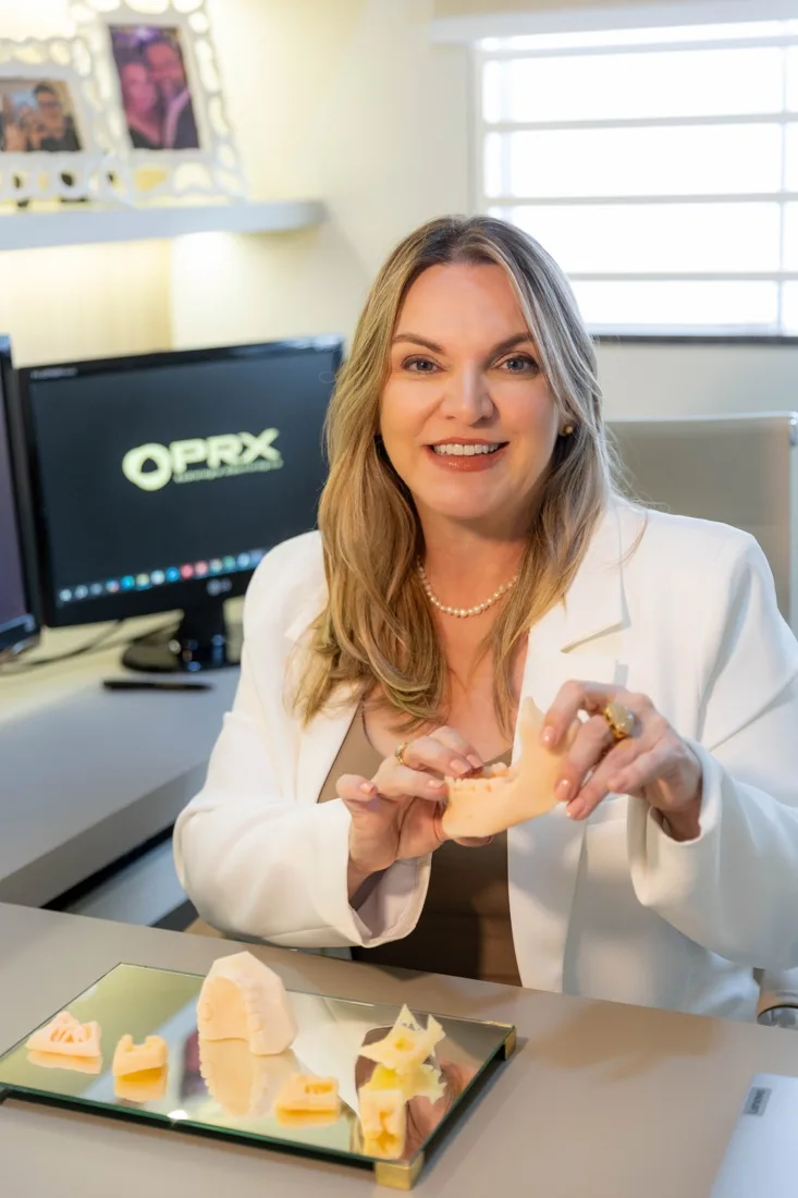 Dra. Fabiana Fernandes Segato segurando um biomodelo de mandíbula impresso em 3D, com outros modelos sobre a mesa